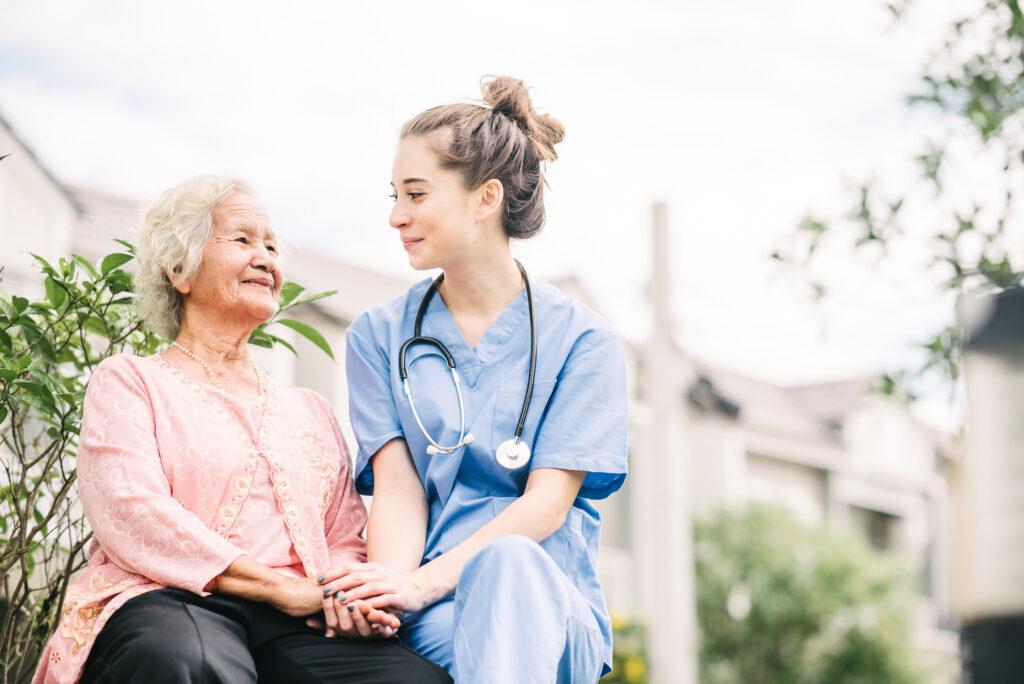 A smiling nurse or caregiver holding the hand of a happy elderly Asian woman, outdoors in a park setting.